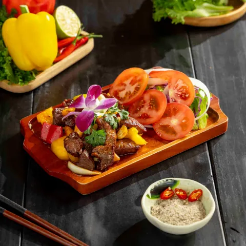 Bò lúc lắc cắt khối xào cùng ớt chuông, bày trên thớt gỗ với cà chua và salad