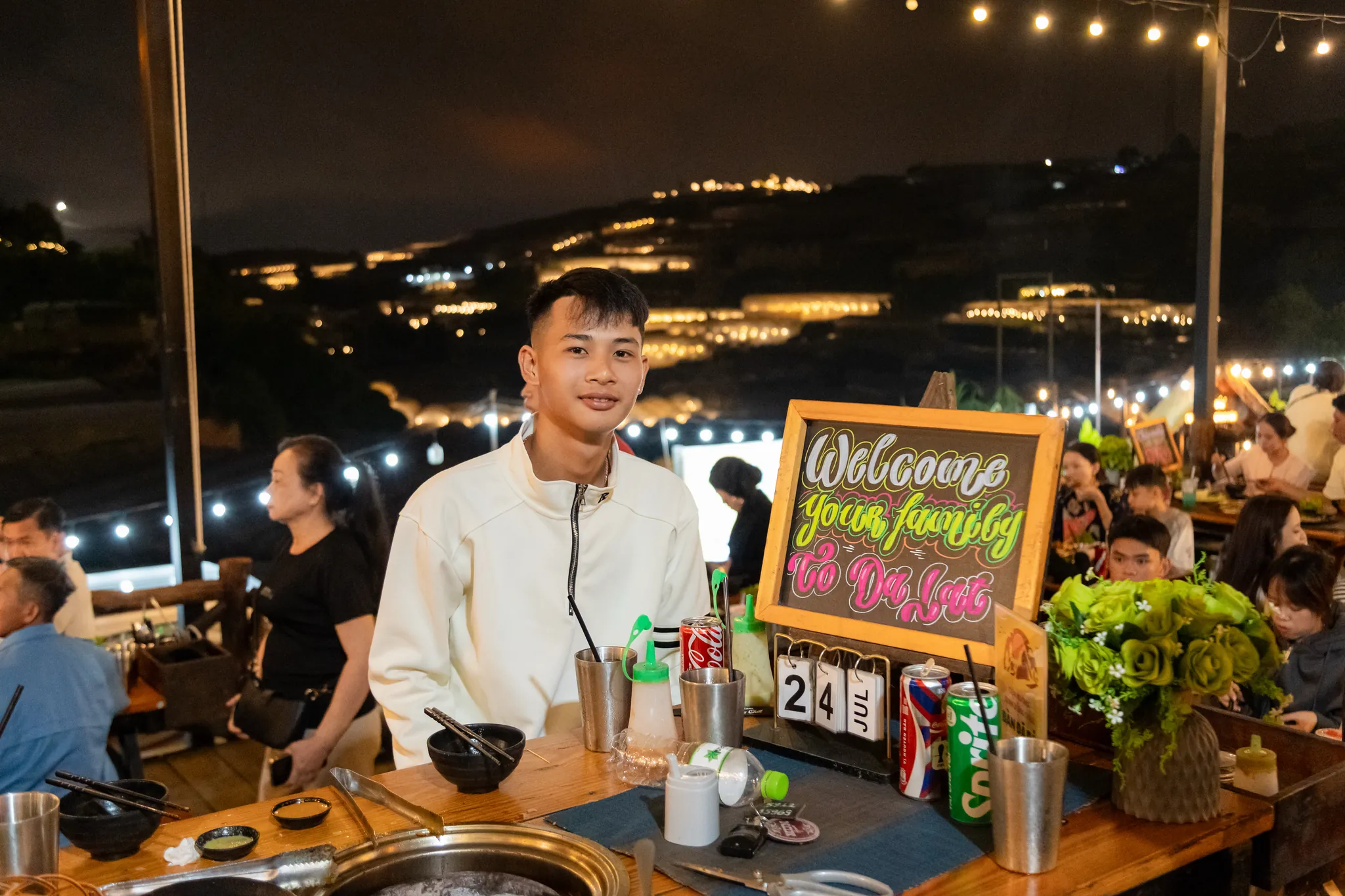 Bảng chào mừng viết tay Welcome your family to Đà Lạt và bộ lịch gỗ trên bàn nướng, khu bàn buổi tối đông khách