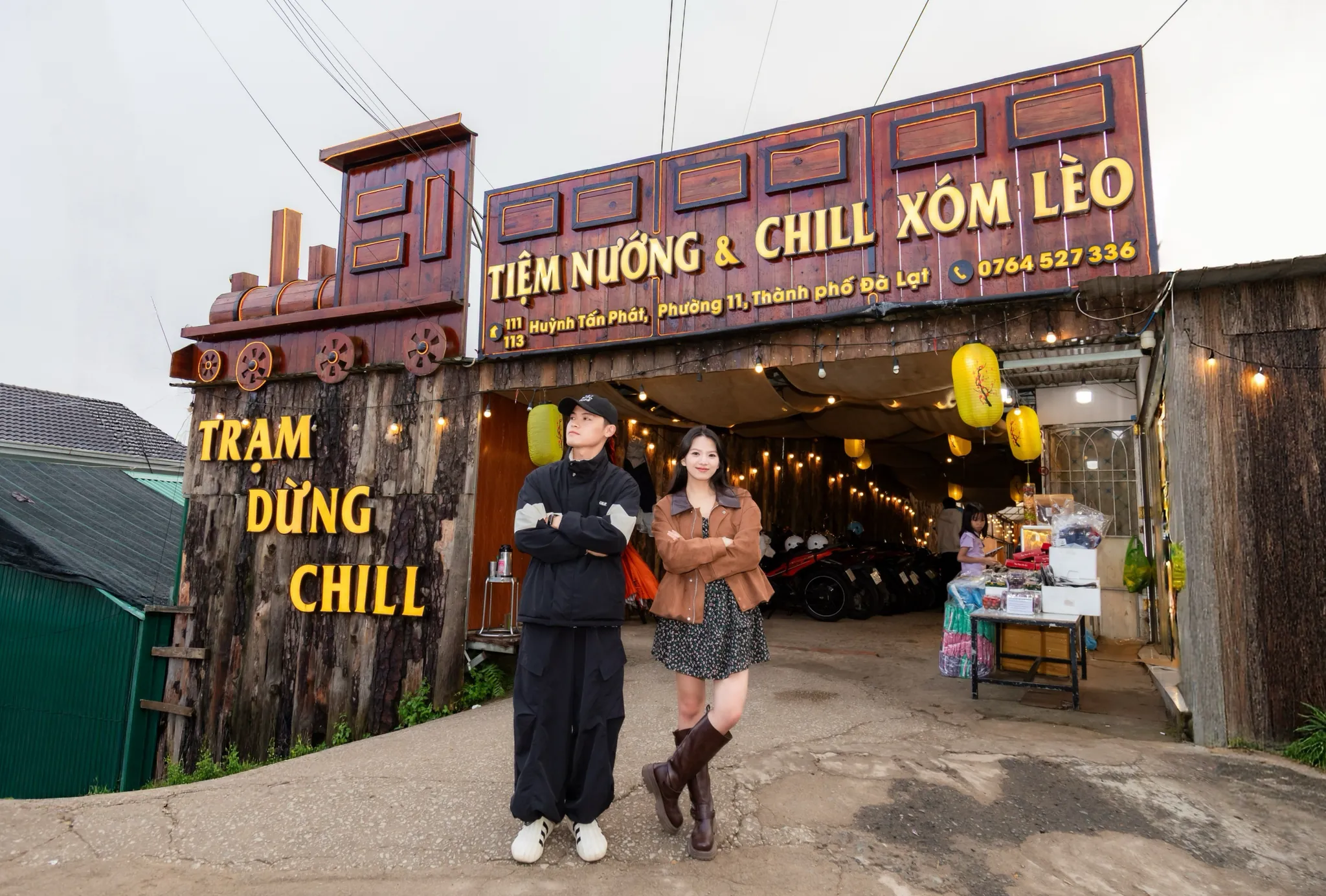 Cặp đôi đứng trước cổng Tiệm Nướng & Chill Xóm Lèo có đầu tàu bằng gỗ và bảng hiệu ghi số 0764 527 336