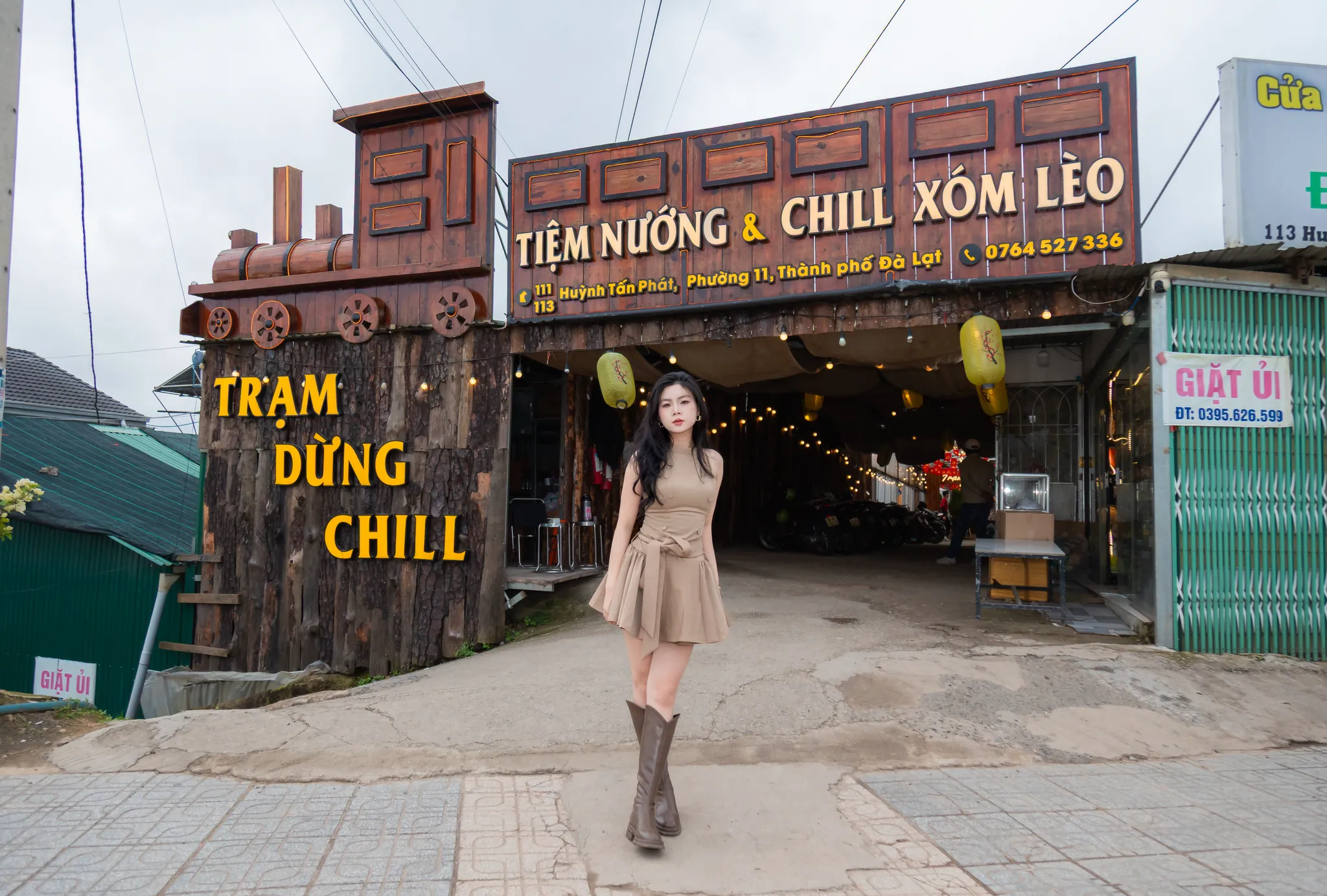 Cô gái đứng trước cổng Tiệm Nướng & Chill Xóm Lèo có đầu tàu bằng gỗ