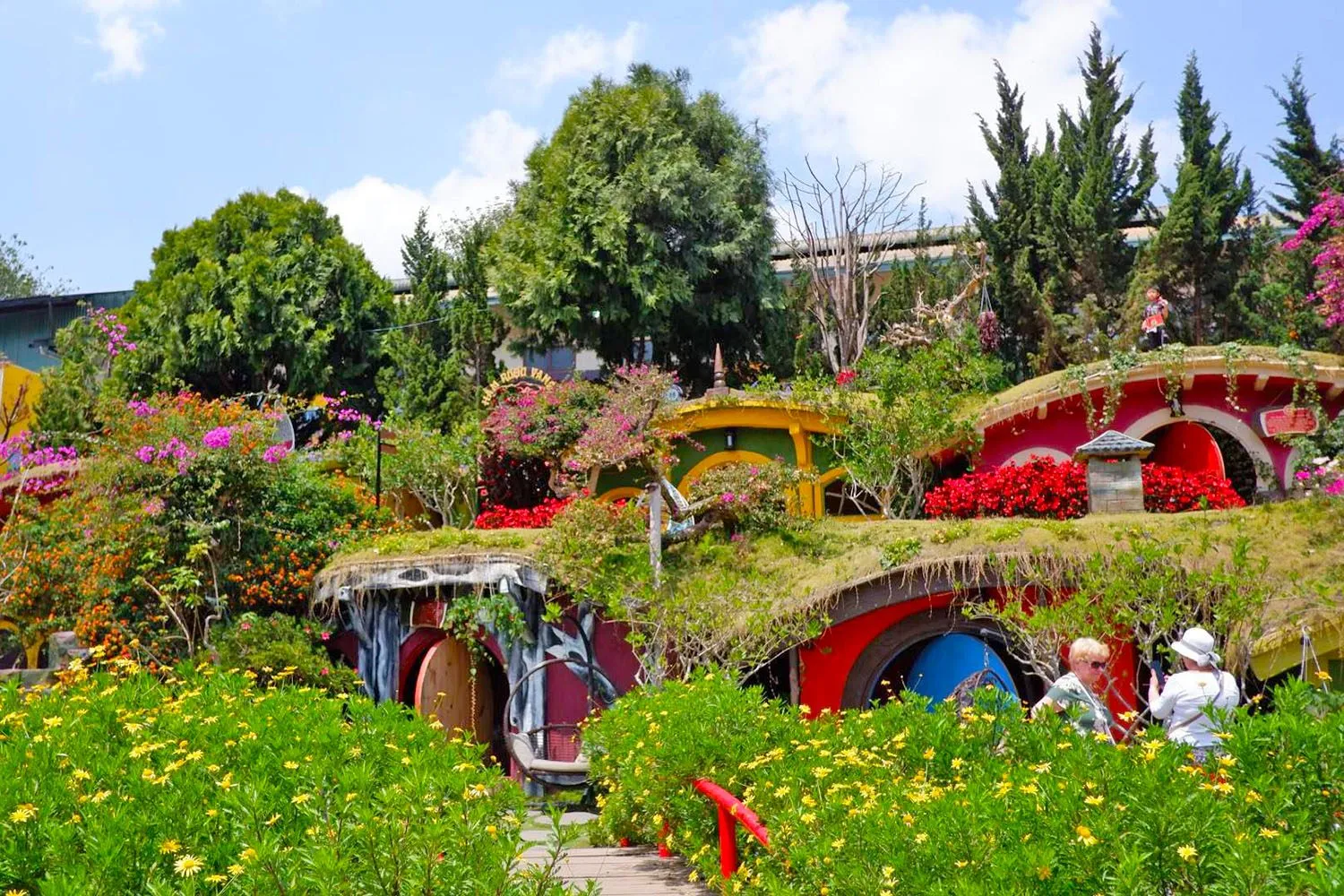 Cụm nhà hobbit mái cỏ sơn đỏ, vàng, xanh nép vào sườn đồi hoa ở Fairytale Land