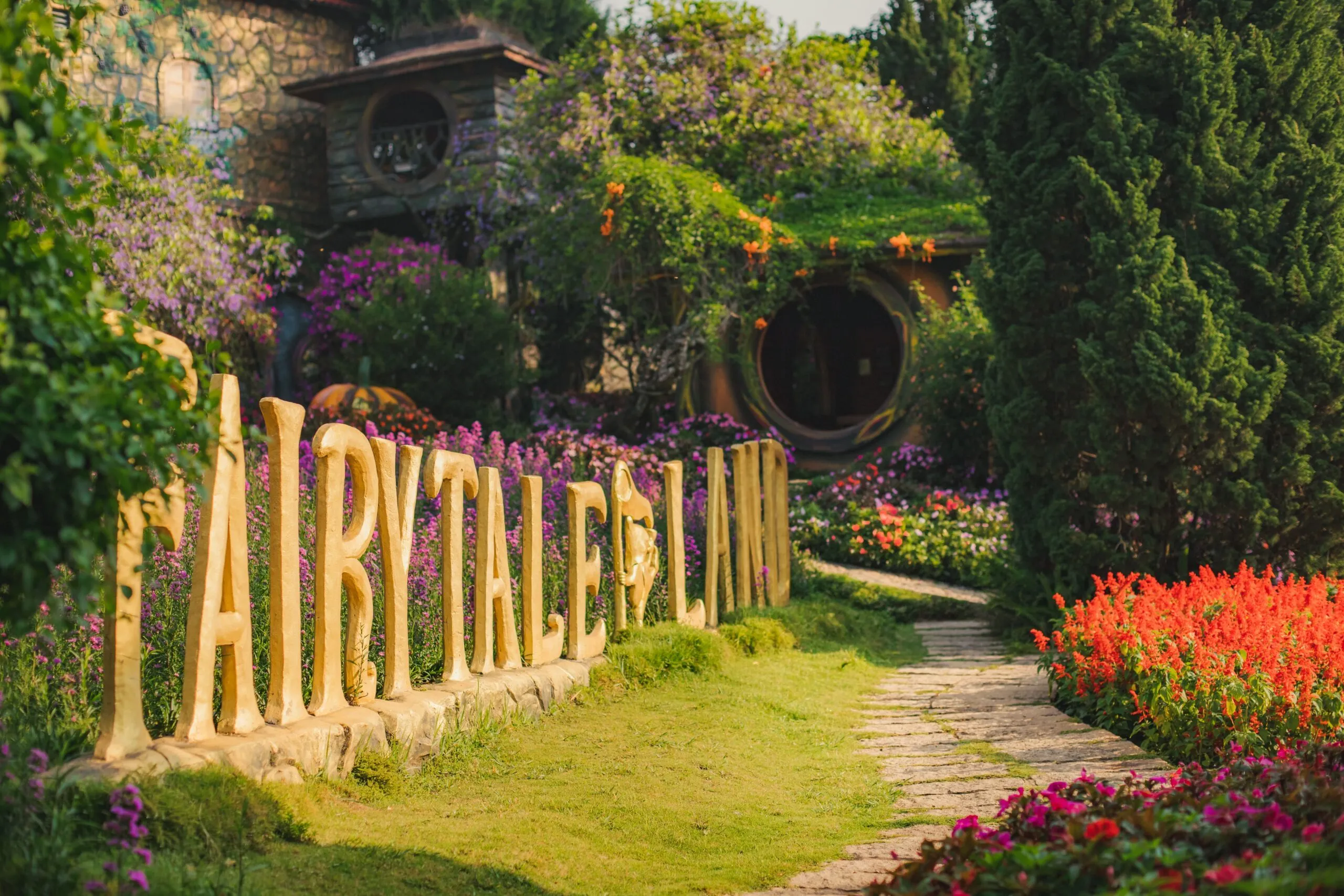 Chữ FAIRYTALE LAND bằng đá đặt giữa luống hoa, lối đi lát đá dẫn vào căn nhà cửa tròn