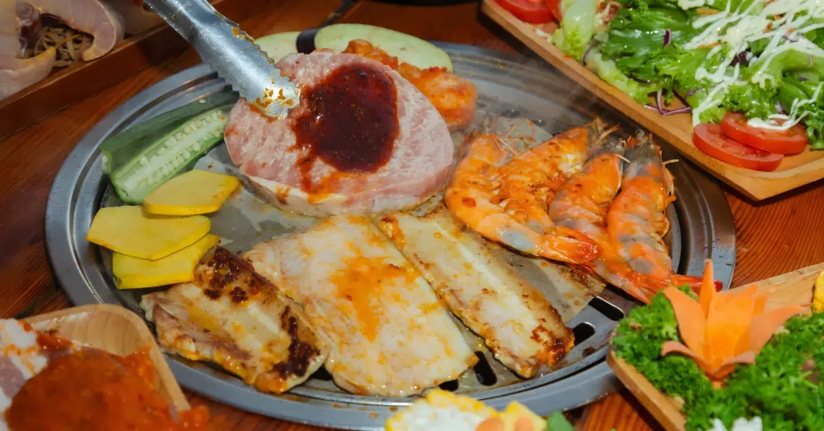 Vỉ nướng với ba chỉ heo sốt, tôm sú, cá phi lê và bí ngòi, xung quanh là đĩa salad và rau sống
