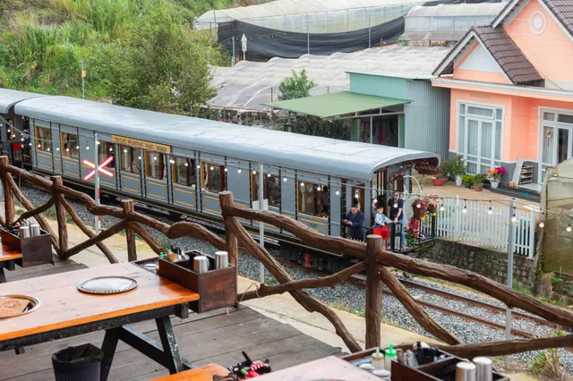 야외 바베큐 테이블의 나무 난간 바로 옆을 지나가는 Dalat Plateau Rail Road 열차, 마지막 객차 문가에 선 승객