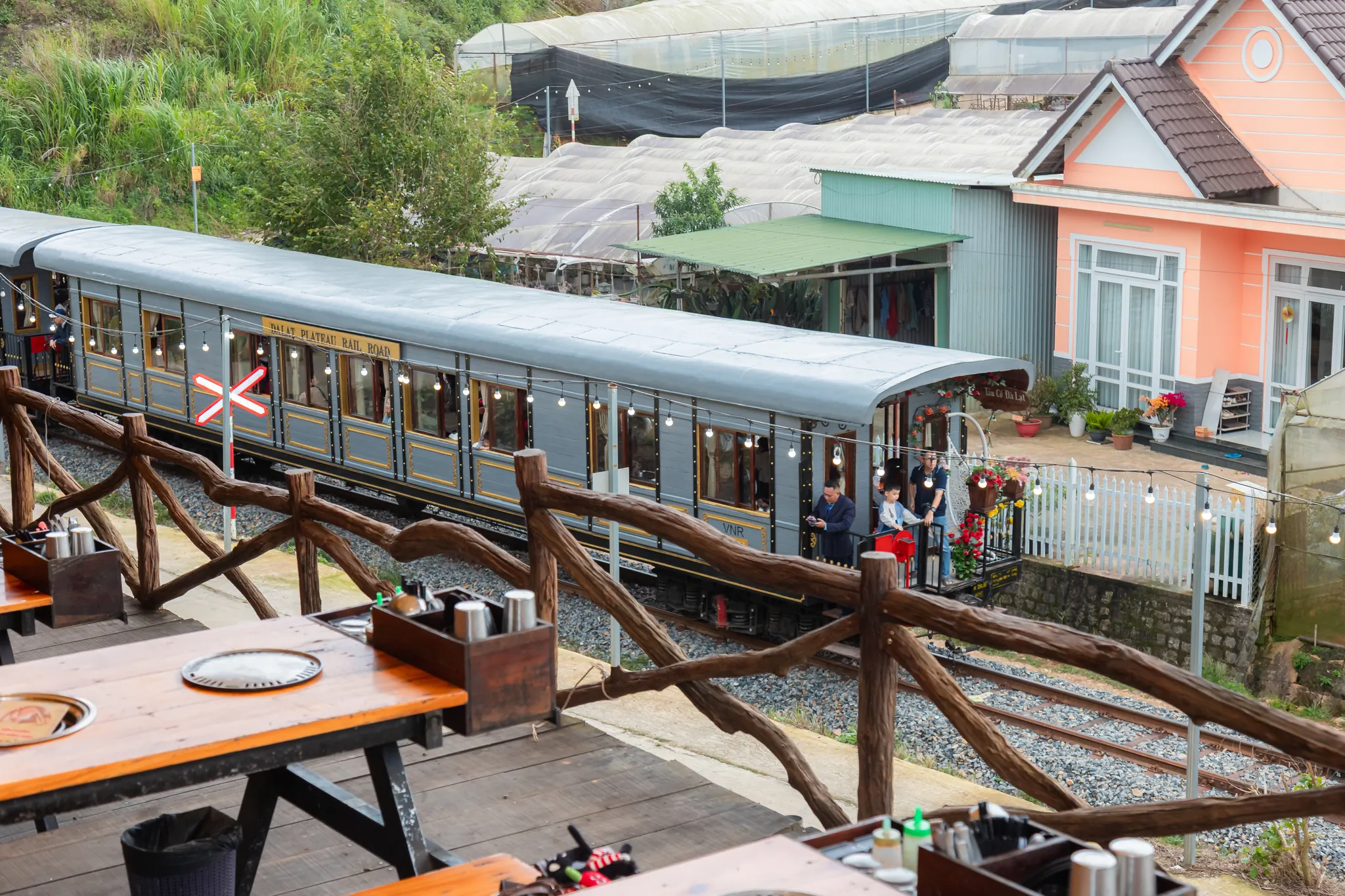 Đoàn tàu Dalat Plateau Rail Road đi ngang sát lan can gỗ của dãy bàn nướng ngoài trời, khách đứng ở cửa toa cuối