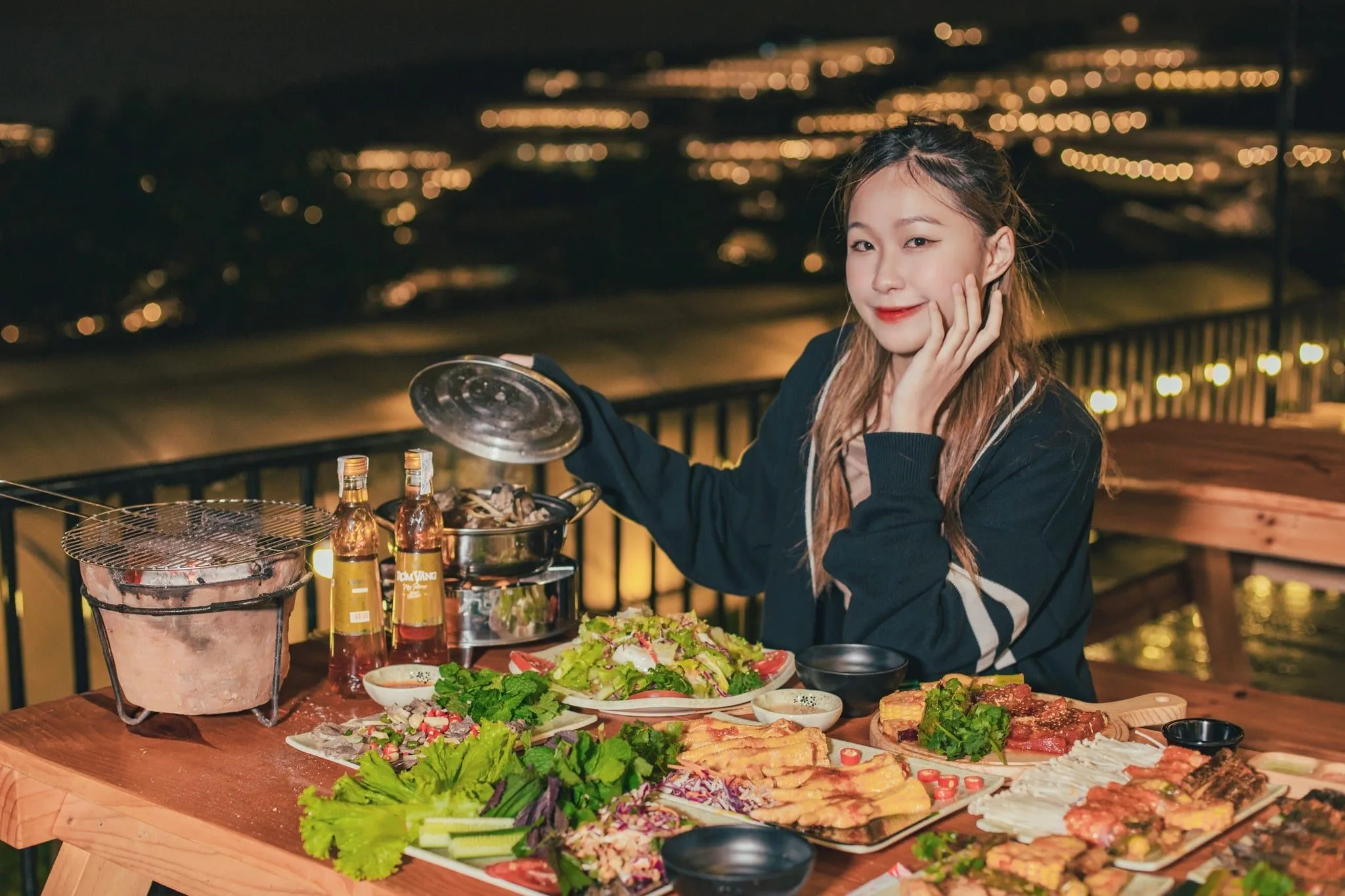 저녁 무렵 Tiệm Nướng & Chill Xóm Lèo의 긴 원목 테이블 위에 놓인 개별 숯불 화로와 전골 냄비, 뒤로 보이는 Trại Mát 계곡의 불빛