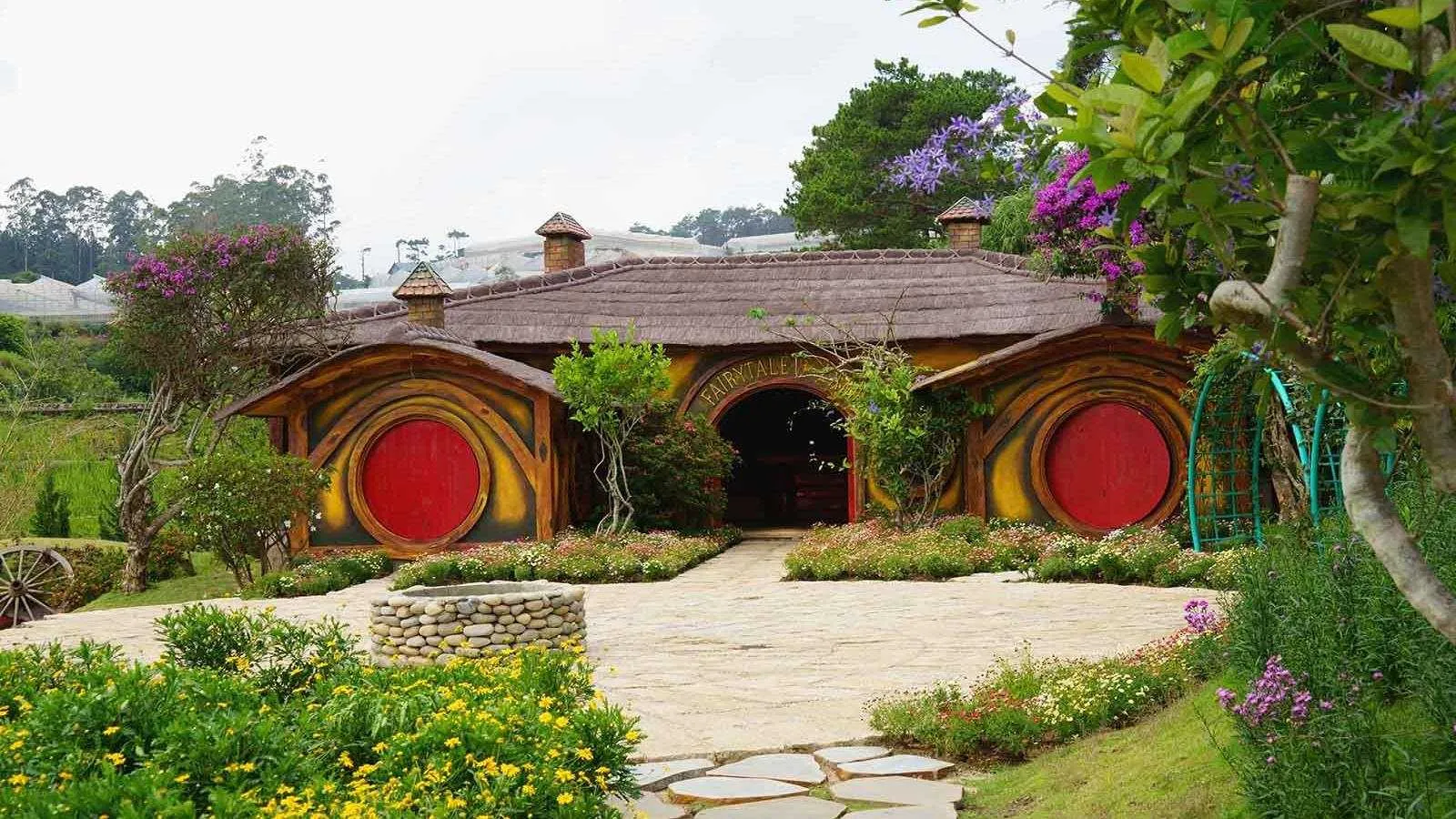 Căn nhà hobbit mái tranh với hai cánh cửa tròn sơn đỏ, lối đi lát đá và giếng đá phía trước