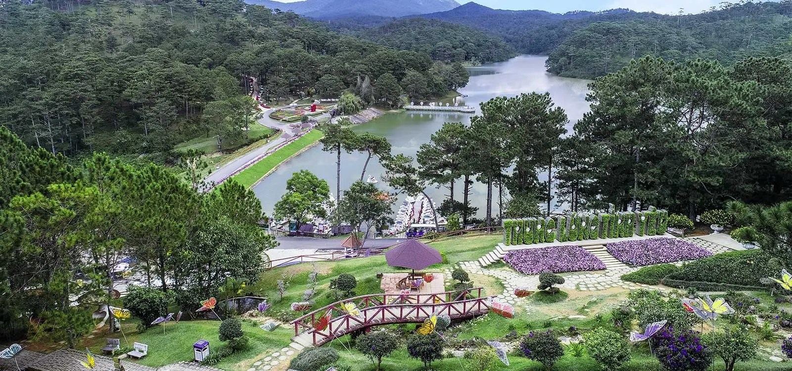 Vườn hoa tím và cầu gỗ đỏ bên hồ Thung Lũng Tình Yêu nhìn từ trên cao, chữ tên khu du lịch kết bằng cây xanh