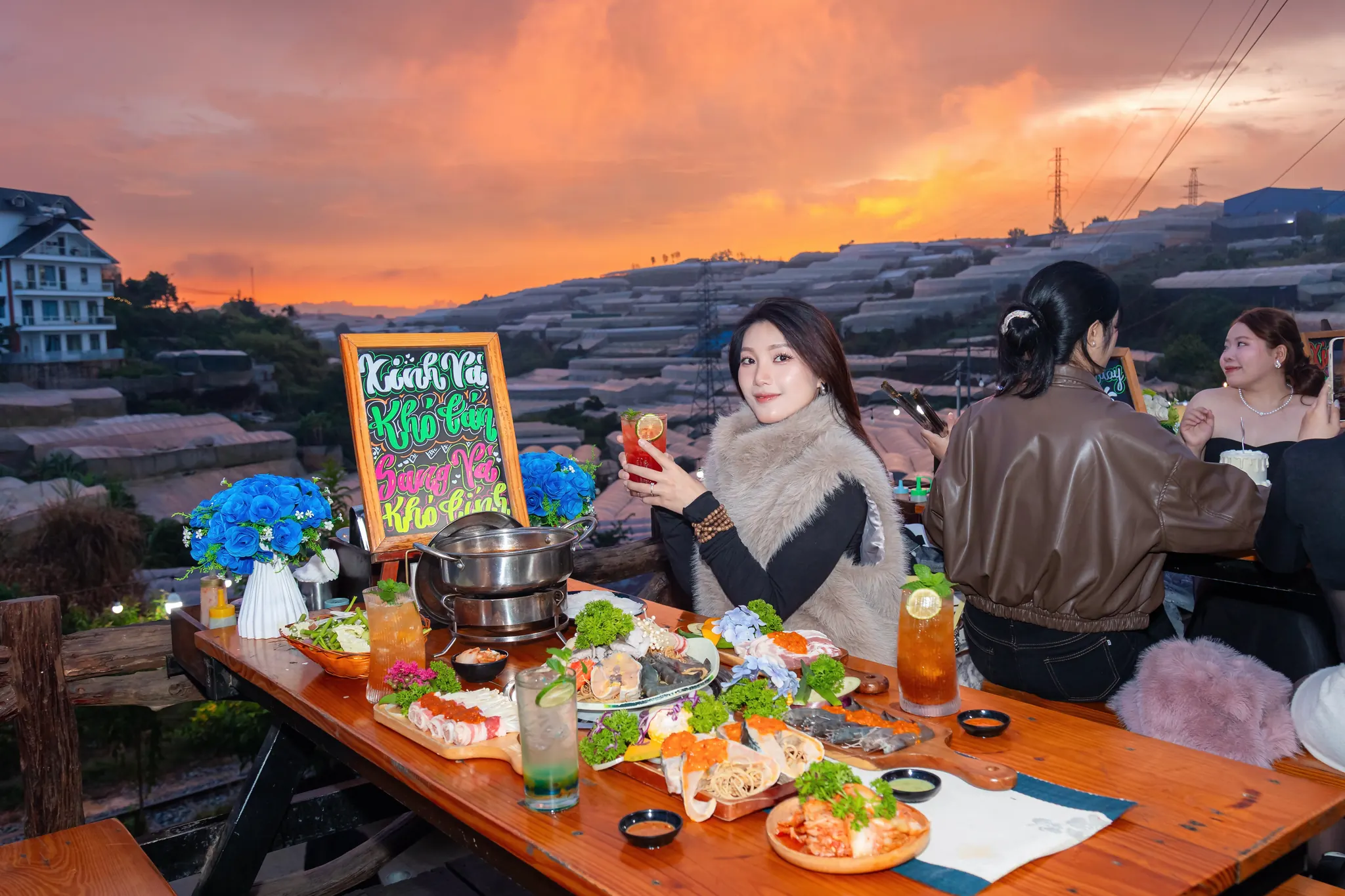 Khách nâng ly bên bàn lẩu nướng lúc hoàng hôn ở Tiệm Nướng & Chill Xóm Lèo, phía sau là thung lũng nhà kính Trại Mát